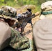 Two individuals in military camouflage uniforms are in a prone shooting position in an outdoor field environment. One person is actively aiming a precision rifle equipped with a telescopic scope and bipod, while the second person observes from close proximity. The setting appears to be a training exercise or range activity based on the controlled positioning.