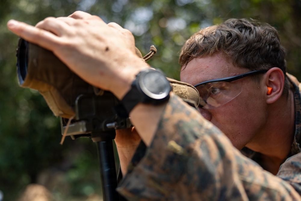 A male service member in MARPAT (Marine pattern) camouflage uniform is looking through what appears to be a spotting scope or rangefinder mounted on a bipod. He is wearing protective eyewear, orange foam earplugs, and a black tactical watch. The setting appears to be an outdoor range or field environment with foliage visible in the background.
