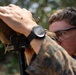 A male service member in MARPAT (Marine pattern) camouflage uniform is looking through what appears to be a spotting scope or rangefinder mounted on a bipod. He is wearing protective eyewear, orange foam earplugs, and a black tactical watch. The setting appears to be an outdoor range or field environment with foliage visible in the background.