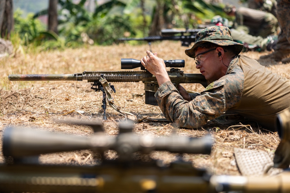 A military service member in MARPAT (Marine pattern) camouflage uniform is in a prone shooting position on dry grass, aiming a precision rifle equipped with a scope and bipod. The setting appears to be an outdoor training range in a tropical or subtropical environment. Additional weapons and personnel are visible in the background and foreground, suggesting a multi-person training exercise.