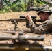 A military service member in MARPAT (Marine pattern) camouflage uniform is in a prone shooting position on dry grass, aiming a precision rifle equipped with a scope and bipod. The setting appears to be an outdoor training range in a tropical or subtropical environment. Additional weapons and personnel are visible in the background and foreground, suggesting a multi-person training exercise.