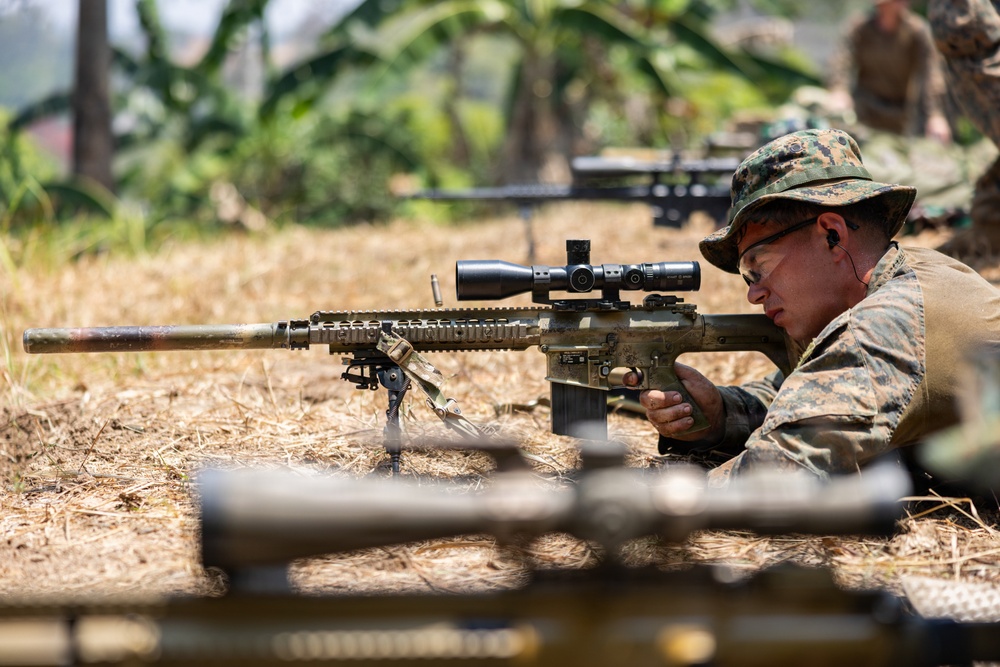 A military sniper in MARPAT (Marine pattern) camouflage uniform is in a prone firing position on dry grass, aiming a scoped precision rifle with a suppressor and bipod. Additional personnel and at least one other sniper rifle are visible in the background. The setting appears to be an outdoor training or operational environment with tropical vegetation.