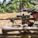 A military sniper in MARPAT (Marine pattern) camouflage uniform is in a prone firing position on dry grass, aiming a scoped precision rifle with a suppressor and bipod. Additional personnel and at least one other sniper rifle are visible in the background. The setting appears to be an outdoor training or operational environment with tropical vegetation.