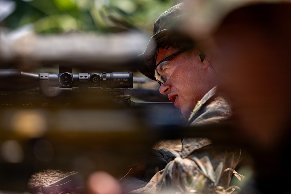 A person wearing camouflage military-style clothing and a boonie hat is positioned behind a rifle equipped with a telescopic scope. The shooter appears to be in a prone or low position, aiming through the optic. The image is captured through foreground foliage, creating a partially obscured, close-up perspective.