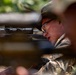 A person wearing camouflage military-style clothing and a boonie hat is positioned behind a rifle equipped with a telescopic scope. The shooter appears to be in a prone or low position, aiming through the optic. The image is captured through foreground foliage, creating a partially obscured, close-up perspective.