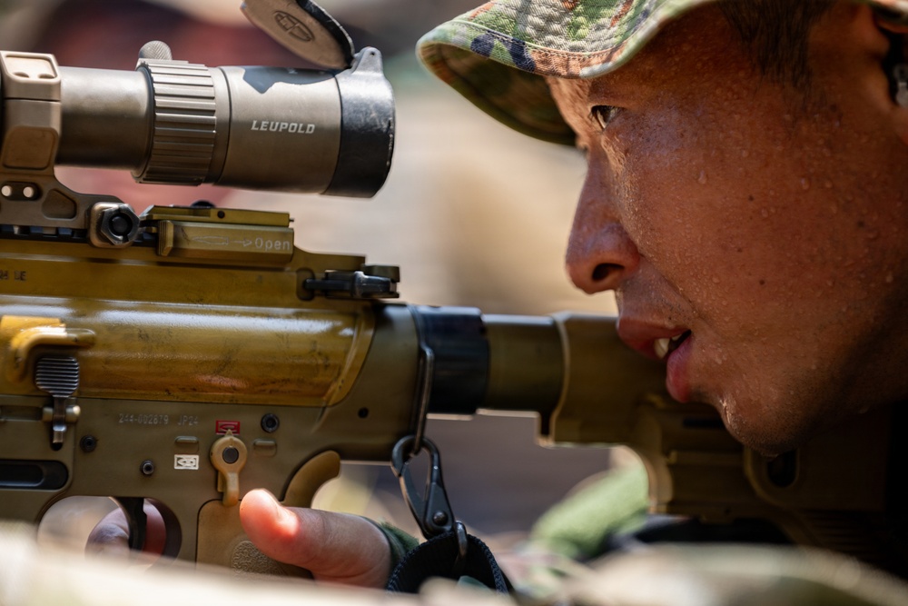 A close-up photograph of a military soldier aiming down the scope of a rifle equipped with a Leupold optic. The soldier is wearing camouflage headgear and appears to be in a prone or low shooting position. The image captures fine detail of both the weapon system and the shooter's focused expression.