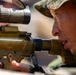 A close-up photograph of a military soldier aiming down the scope of a rifle equipped with a Leupold optic. The soldier is wearing camouflage headgear and appears to be in a prone or low shooting position. The image captures fine detail of both the weapon system and the shooter's focused expression.