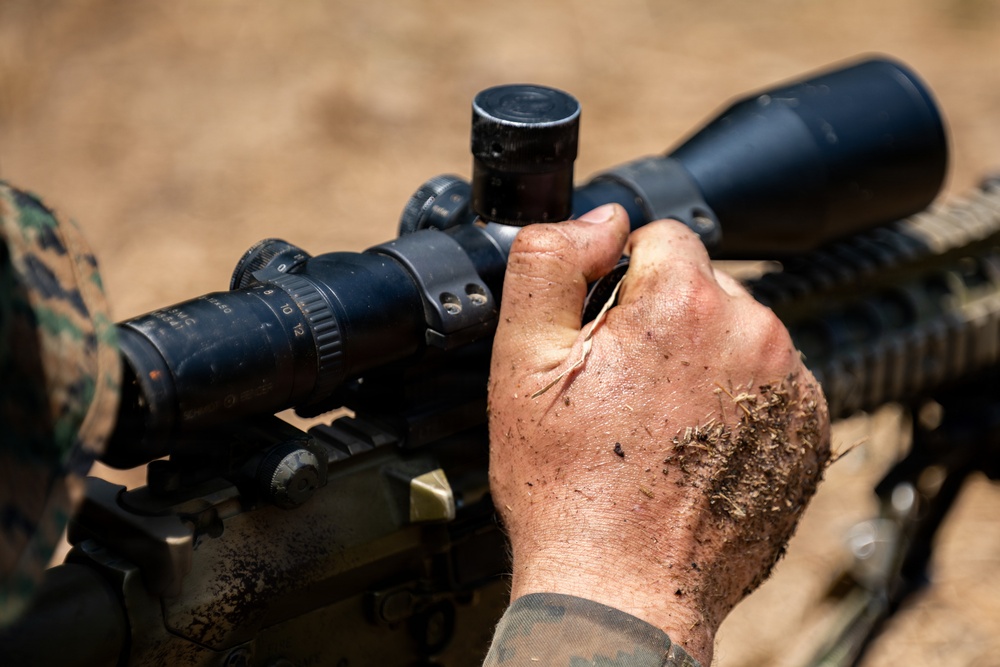 A close-up image showing a person in military camouflage uniform adjusting or manipulating a rifle scope mounted on a firearm. The individual's hands are visibly dirty and covered in soil and debris, suggesting field conditions. The focus is on the tactical optic and the adjustment being made to its turrets or settings.