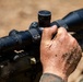 A close-up image showing a person in military camouflage uniform adjusting or manipulating a rifle scope mounted on a firearm. The individual's hands are visibly dirty and covered in soil and debris, suggesting field conditions. The focus is on the tactical optic and the adjustment being made to its turrets or settings.