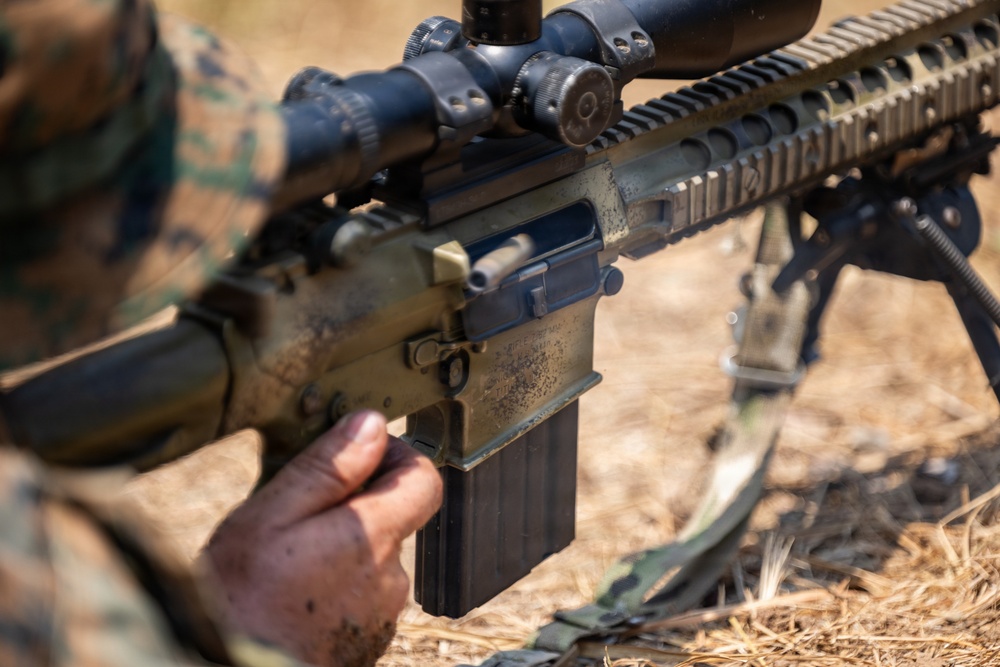 A military service member in woodland camouflage uniform is handling a scoped semi-automatic rifle, appearing to insert or remove a magazine. The weapon is equipped with a magnified optic scope, Picatinny rail system, and a bipod, suggesting a designated marksman or sniper configuration. The setting appears to be an outdoor training or field environment with dry grass visible in the background.