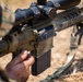 A military service member in woodland camouflage uniform is handling a scoped semi-automatic rifle, appearing to insert or remove a magazine. The weapon is equipped with a magnified optic scope, Picatinny rail system, and a bipod, suggesting a designated marksman or sniper configuration. The setting appears to be an outdoor training or field environment with dry grass visible in the background.