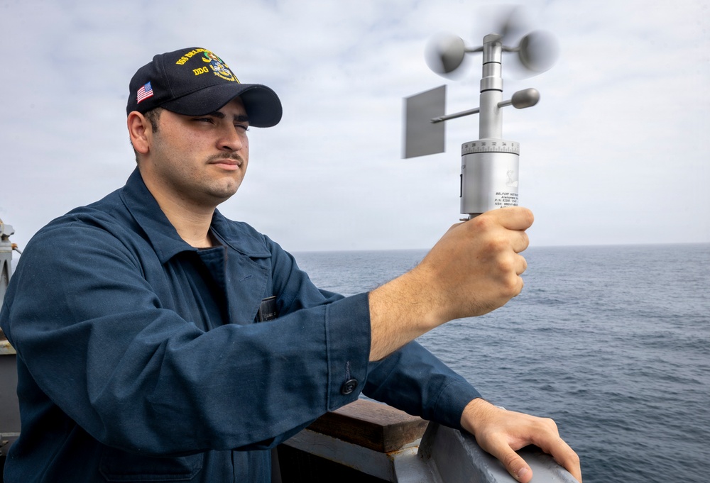 A U.S. Navy sailor aboard a DDG (destroyer) is holding a handheld anemometer to measure wind speed and direction while standing on the ship's deck. The sailor is wearing standard Navy blue coveralls and a DDG-designated ball cap with an American flag patch. The ocean is visible in the background under an overcast sky.