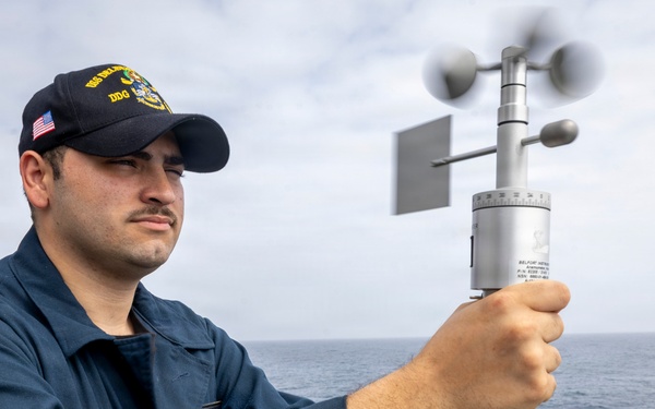 A U.S. Navy sailor aboard a DDG (destroyer) is holding a handheld anemometer to measure wind speed and direction while standing on the ship's deck. The sailor is wearing standard Navy blue coveralls and a DDG-designated ball cap with an American flag patch. The ocean is visible in the background under an overcast sky.