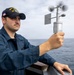 A U.S. Navy sailor aboard a DDG (destroyer) is holding a handheld anemometer to measure wind speed and direction while standing on the ship's deck. The sailor is wearing standard Navy blue coveralls and a DDG-designated ball cap with an American flag patch. The ocean is visible in the background under an overcast sky.