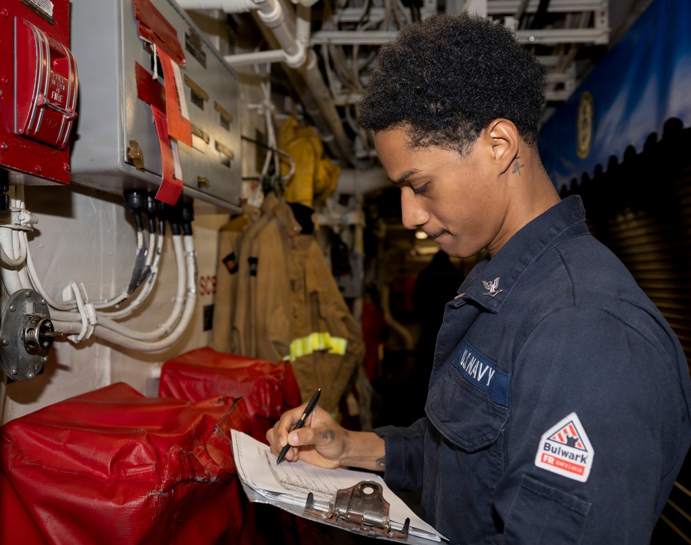 A U.S. Navy sailor in dark blue coveralls is writing on a clipboard in what appears to be a shipboard compartment or passageway. The individual is conducting what appears to be an inspection or documentation task near fire safety equipment. The setting is consistent with the interior of a U.S. Navy vessel.