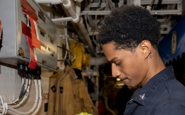 A U.S. Navy sailor in dark blue coveralls is writing on a clipboard in what appears to be a shipboard compartment or passageway. The individual is conducting what appears to be an inspection or documentation task near fire safety equipment. The setting is consistent with the interior of a U.S. Navy vessel.