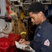 A U.S. Navy sailor in dark blue coveralls is writing on a clipboard in what appears to be a shipboard compartment or passageway. The individual is conducting what appears to be an inspection or documentation task near fire safety equipment. The setting is consistent with the interior of a U.S. Navy vessel.