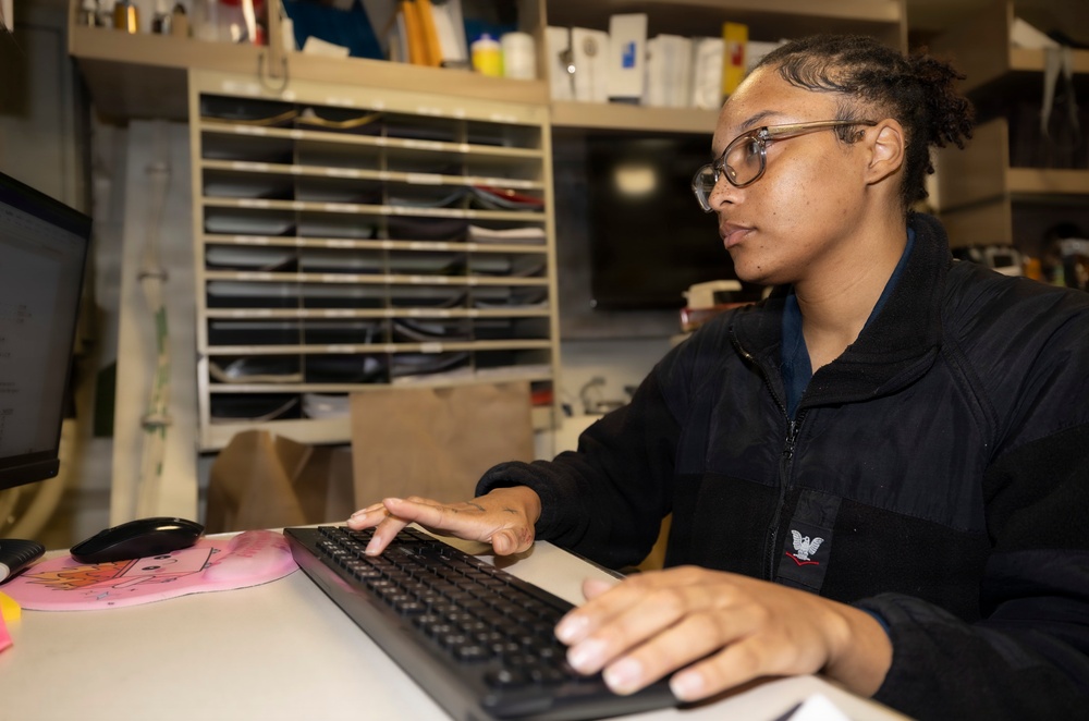 A young person wearing glasses and a black zip-up jacket with an eagle emblem is seated at a desk working on a computer. They are typing on a black keyboard while looking at a monitor to their left. The workspace appears to be a busy office or administrative area with filing shelves and various supplies visible in the background.