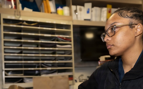 A young person wearing glasses and a black zip-up jacket with an eagle emblem is seated at a desk working on a computer. They are typing on a black keyboard while looking at a monitor to their left. The workspace appears to be a busy office or administrative area with filing shelves and various supplies visible in the background.