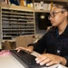 A young person wearing glasses and a black zip-up jacket with an eagle emblem is seated at a desk working on a computer. They are typing on a black keyboard while looking at a monitor to their left. The workspace appears to be a busy office or administrative area with filing shelves and various supplies visible in the background.
