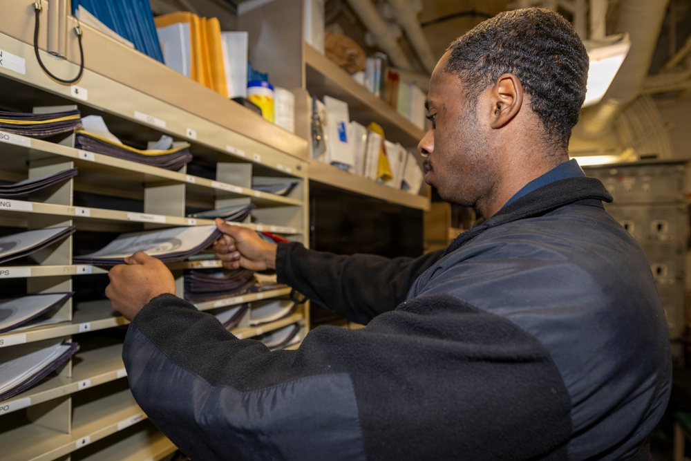 A man wearing a dark navy fleece jacket is sorting or retrieving documents from a multi-slot mail/filing organizer unit. The setting appears to be a compact storage or mailroom space, possibly aboard a naval vessel given the visible structural elements in the background. The shelving unit contains numerous documents, folders, and papers organized across multiple labeled compartments.