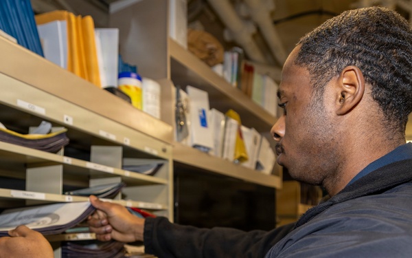 A man wearing a dark navy fleece jacket is sorting or retrieving documents from a multi-slot mail/filing organizer unit. The setting appears to be a compact storage or mailroom space, possibly aboard a naval vessel given the visible structural elements in the background. The shelving unit contains numerous documents, folders, and papers organized across multiple labeled compartments.