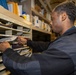 A man wearing a dark navy fleece jacket is sorting or retrieving documents from a multi-slot mail/filing organizer unit. The setting appears to be a compact storage or mailroom space, possibly aboard a naval vessel given the visible structural elements in the background. The shelving unit contains numerous documents, folders, and papers organized across multiple labeled compartments.