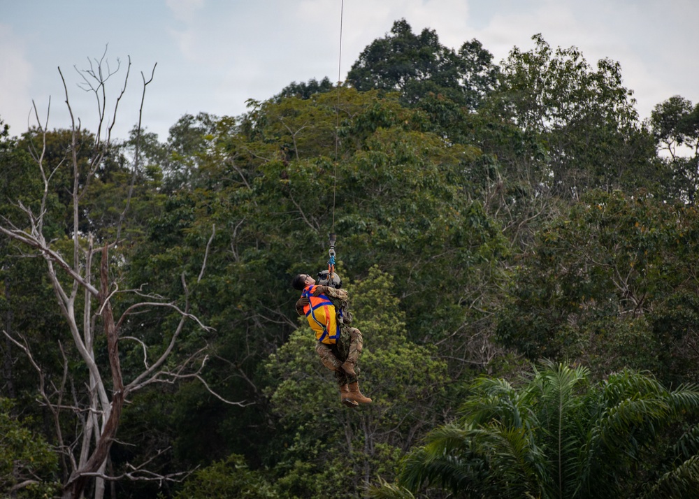 A military or rescue personnel in camouflage uniform is being hoisted by a cable/hoist system while carrying or securing a person wearing a bright orange and yellow life vest or rescue harness. The operation appears to be a helicopter hoist rescue or training exercise conducted over a tropical forested area. The rescuer is looking upward toward the cable attachment point while suspended mid-air.