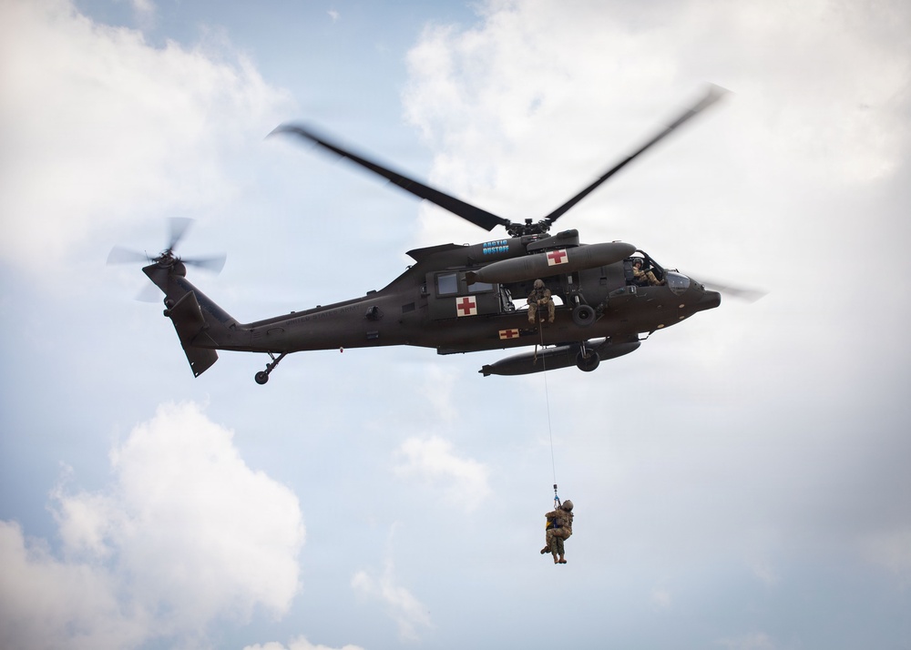 A military medical evacuation (MEDEVAC) helicopter, identifiable by multiple red cross markings, is airborne and conducting a hoist rescue operation. A person in military gear is suspended below the aircraft on a hoist cable, being lifted toward the open side door where crew members are visible. The scene appears to be a training exercise or active rescue operation against a partly cloudy sky.