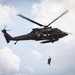 A military medical evacuation (MEDEVAC) helicopter, identifiable by multiple red cross markings, is airborne and conducting a hoist rescue operation. A person in military gear is suspended below the aircraft on a hoist cable, being lifted toward the open side door where crew members are visible. The scene appears to be a training exercise or active rescue operation against a partly cloudy sky.
