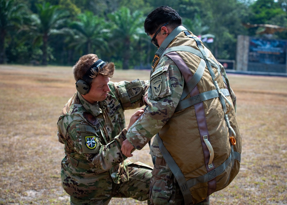 Two military personnel in OCP (Operational Camouflage Pattern) uniforms are conducting a pre-jump equipment inspection on a grassy field surrounded by tropical vegetation. One soldier, wearing hearing protection, is kneeling and checking the parachute harness and equipment of a standing soldier who is wearing a full parachute rig. The setting and patches suggest a U.S. Army airborne operation, likely during a joint or multinational exercise.