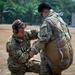 Two military personnel in OCP (Operational Camouflage Pattern) uniforms are conducting a pre-jump equipment inspection on a grassy field surrounded by tropical vegetation. One soldier, wearing hearing protection, is kneeling and checking the parachute harness and equipment of a standing soldier who is wearing a full parachute rig. The setting and patches suggest a U.S. Army airborne operation, likely during a joint or multinational exercise.