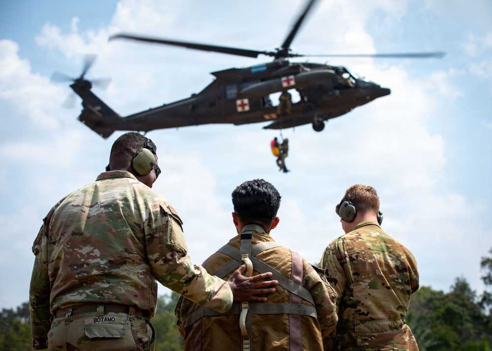 Three military personnel in camouflage uniforms observe a medical evacuation (MEDEVAC) helicopter hoist operation. A UH-60 Black Hawk helicopter marked with a red cross is hovering overhead while a person is being hoisted on a cable. The soldier on the left, identified by name tape as 'BOTAMO,' has his hand on the middle soldier's back.