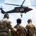 Three military personnel in camouflage uniforms observe a medical evacuation (MEDEVAC) helicopter hoist operation. A UH-60 Black Hawk helicopter marked with a red cross is hovering overhead while a person is being hoisted on a cable. The soldier on the left, identified by name tape as 'BOTAMO,' has his hand on the middle soldier's back.