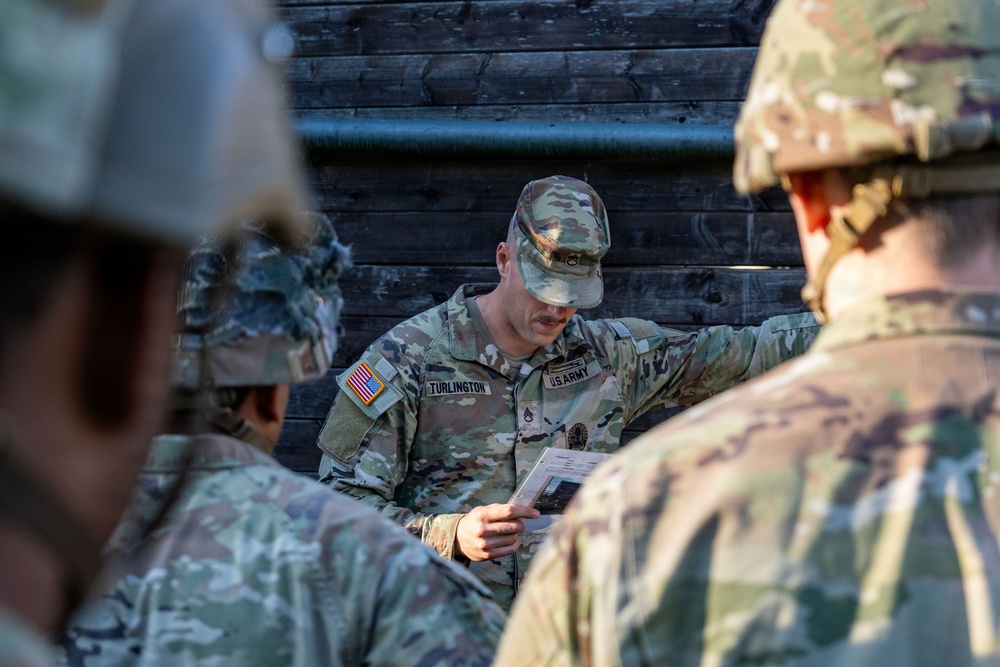 A U.S. Army soldier identified by name tape as 'Turlington' is conducting a briefing or instruction session with other soldiers in military camouflage uniforms. He is holding what appears to be a document or card while gesturing, suggesting he is explaining a task or procedure. The setting appears to be an outdoor training environment with a wooden structure in the background.