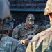 A U.S. Army soldier identified by name tape as 'Turlington' is conducting a briefing or instruction session with other soldiers in military camouflage uniforms. He is holding what appears to be a document or card while gesturing, suggesting he is explaining a task or procedure. The setting appears to be an outdoor training environment with a wooden structure in the background.