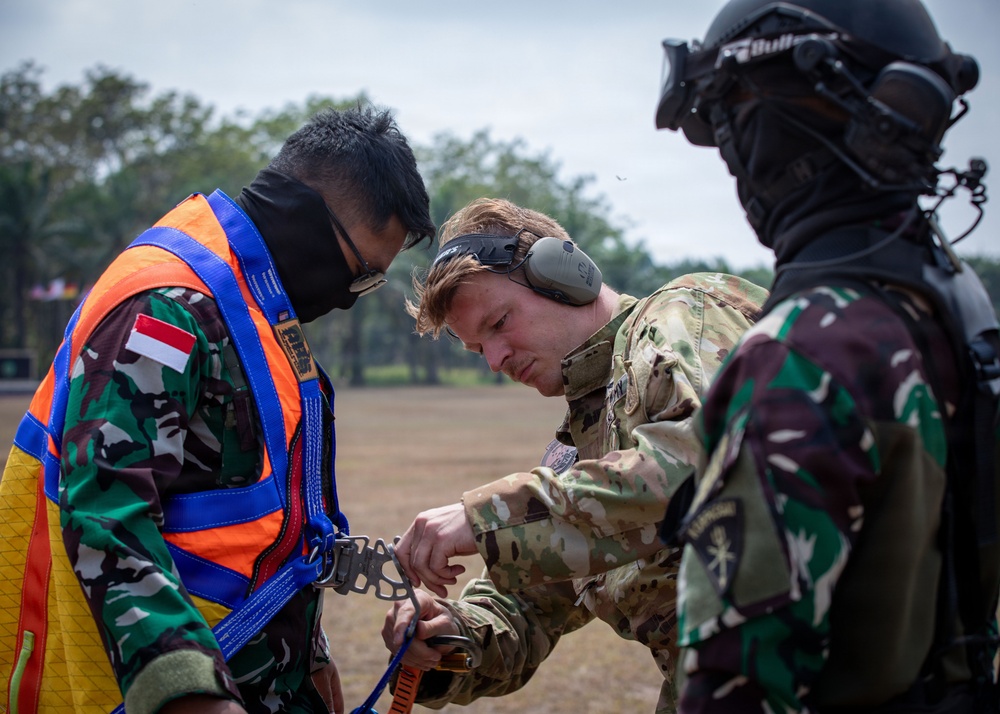 Three military personnel are engaged in what appears to be a pre-jump or equipment check during a joint military exercise. A Western soldier in MultiCam uniform with hearing protection is inspecting or adjusting harness equipment on an Indonesian soldier wearing an orange safety vest over camouflage uniform with an Indonesian flag patch. A third soldier in full tactical gear including helmet and goggles observes from the right.