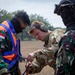 Three military personnel are engaged in what appears to be a pre-jump or equipment check during a joint military exercise. A Western soldier in MultiCam uniform with hearing protection is inspecting or adjusting harness equipment on an Indonesian soldier wearing an orange safety vest over camouflage uniform with an Indonesian flag patch. A third soldier in full tactical gear including helmet and goggles observes from the right.