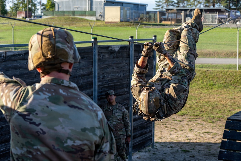Military personnel are conducting a physical training exercise at what appears to be an obstacle course on a military installation. One soldier is traversing a rope or cable obstacle in an inverted position while others observe. The setting includes wooden barriers, metal frame structures, and military buildings in the background.