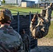 Military personnel are conducting a physical training exercise at what appears to be an obstacle course on a military installation. One soldier is traversing a rope or cable obstacle in an inverted position while others observe. The setting includes wooden barriers, metal frame structures, and military buildings in the background.