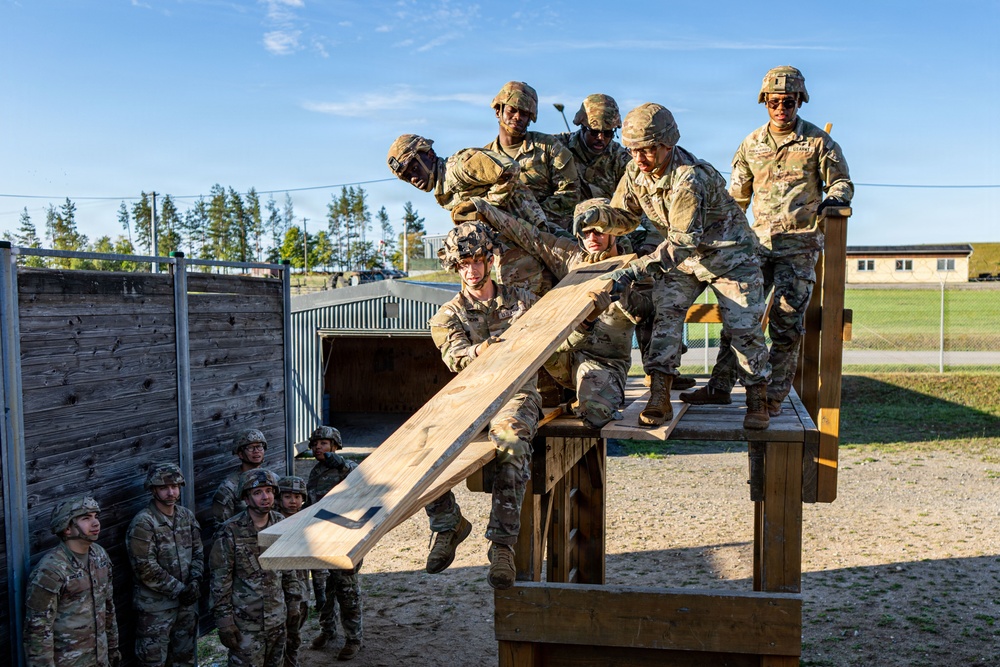 A group of military personnel in camouflage uniforms and helmets are engaged in a team-building obstacle course exercise at what appears to be a military training facility. Several soldiers on an elevated wooden platform are maneuvering a large wooden plank, while a larger group observes from the ground. The setting includes a fenced military compound with trees and buildings visible in the background.