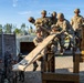 A group of military personnel in camouflage uniforms and helmets are engaged in a team-building obstacle course exercise at what appears to be a military training facility. Several soldiers on an elevated wooden platform are maneuvering a large wooden plank, while a larger group observes from the ground. The setting includes a fenced military compound with trees and buildings visible in the background.