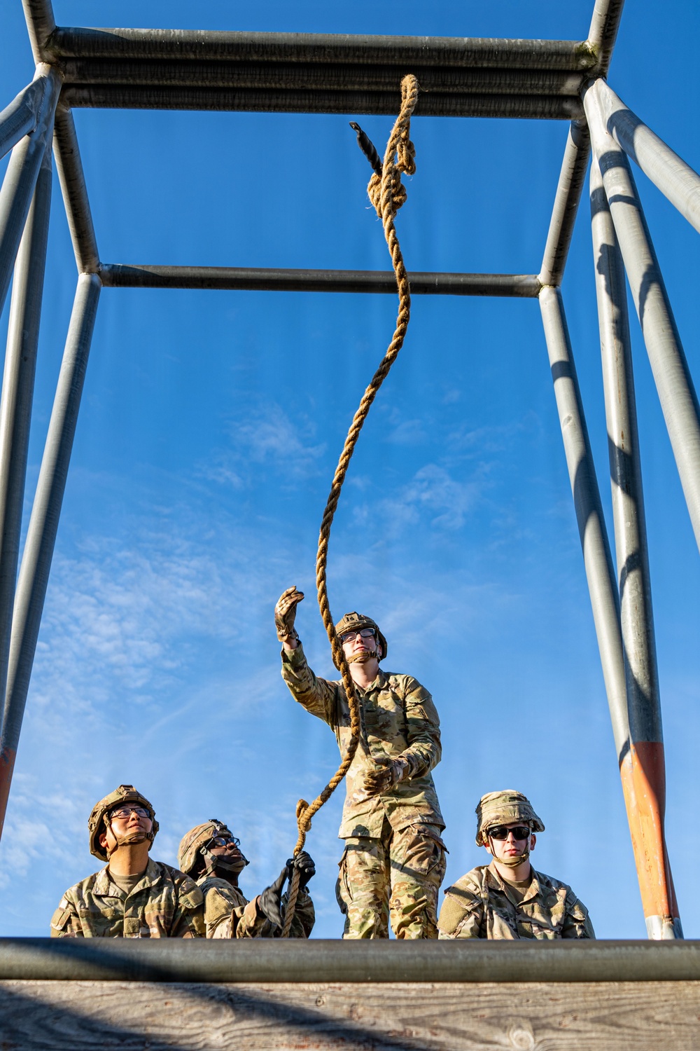 Four military personnel in camouflage uniforms are gathered beneath a metal training structure, with one individual reaching upward to handle a thick rope suspended from the overhead frame. The low-angle perspective looking upward emphasizes the height of the structure against a clear blue sky. This appears to be a military obstacle course or rappelling training exercise.