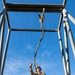 Four military personnel in camouflage uniforms are gathered beneath a metal training structure, with one individual reaching upward to handle a thick rope suspended from the overhead frame. The low-angle perspective looking upward emphasizes the height of the structure against a clear blue sky. This appears to be a military obstacle course or rappelling training exercise.