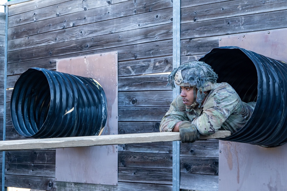 A soldier in full camouflage uniform and combat helmet is emerging from a large black corrugated tunnel mounted on a weathered wooden wall structure. The image appears to capture a military obstacle course or training exercise. The soldier is leaning on a wooden beam as they exit the tunnel opening.