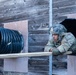 A soldier in full camouflage uniform and combat helmet is emerging from a large black corrugated tunnel mounted on a weathered wooden wall structure. The image appears to capture a military obstacle course or training exercise. The soldier is leaning on a wooden beam as they exit the tunnel opening.