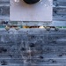 Several people are peering over the top edge of a weathered gray wooden wall or fence, with only their eyes and gloved hands visible above the plank. The image is shot straight-on, creating a symmetrical, humorous composition. A metal panel is mounted on the upper portion of the wooden structure.
