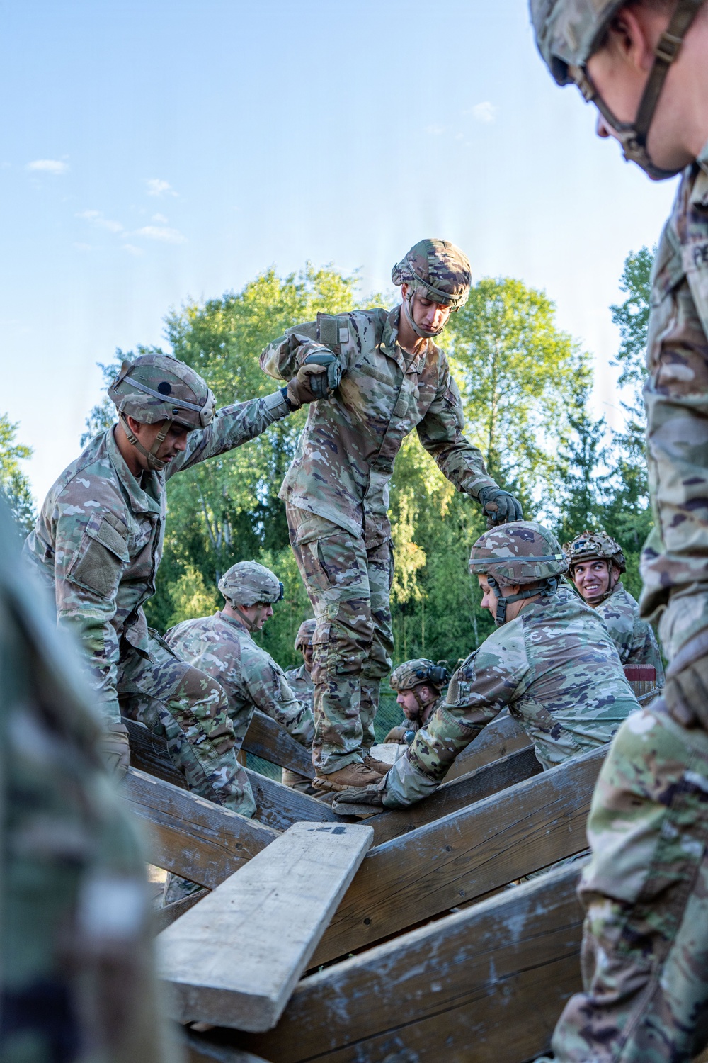 A group of military personnel in MultiCam/OCP camouflage uniforms are engaged in what appears to be a team-building or obstacle course exercise involving wooden planks or beams. One soldier is standing elevated on the wooden structure while others support or stabilize it from below and around the sides. The setting is an outdoor training environment with trees visible in the background under a clear sky.