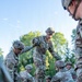 A group of military personnel in MultiCam/OCP camouflage uniforms are engaged in what appears to be a team-building or obstacle course exercise involving wooden planks or beams. One soldier is standing elevated on the wooden structure while others support or stabilize it from below and around the sides. The setting is an outdoor training environment with trees visible in the background under a clear sky.