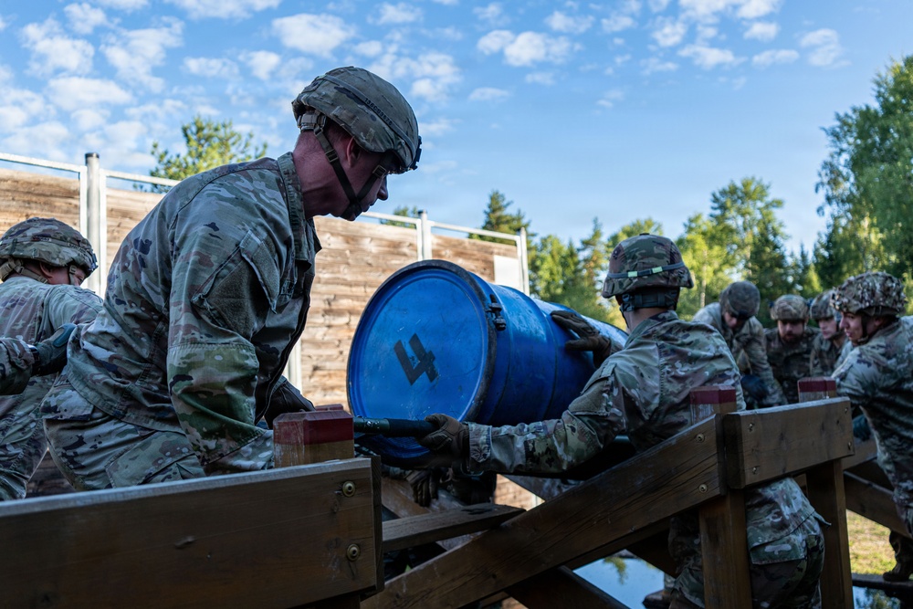 A group of military personnel in camouflage uniforms and helmets are working together to maneuver a blue barrel marked with the number '4' across a wooden structure, likely as part of a team-building or obstacle course exercise. The setting appears to be an outdoor military training area with wooden barriers and lush green trees in the background. The activity demonstrates coordinated teamwork under a training scenario.